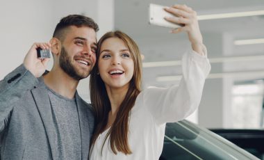 A young couple is standing near the car, and the guy holds the key in hand and the girl holds a mobile and is taking a selfie.